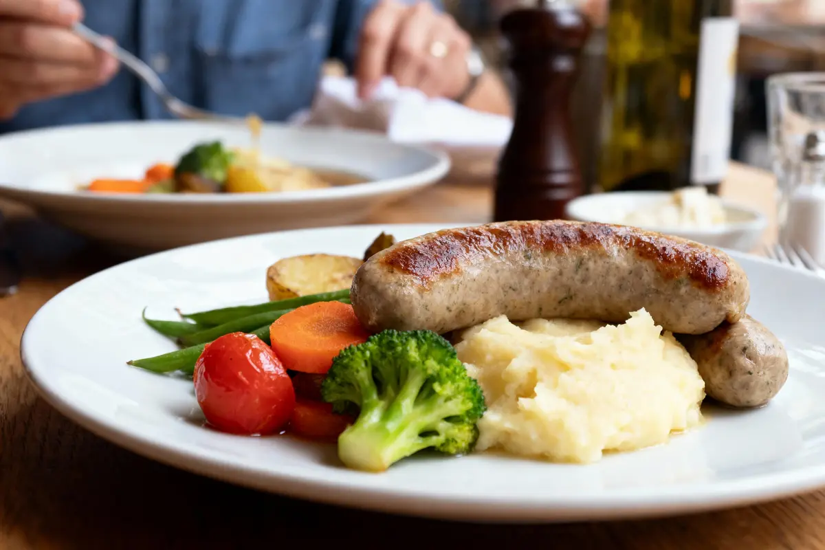 Assiette de saucisses, purée, légumes variés, sur table en bois, homme mangeant en arrière-plan flou.