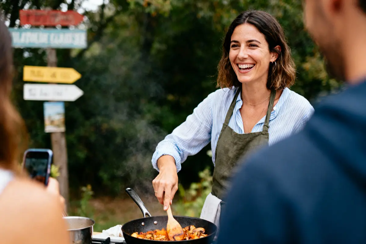 Femme souriante cuisinant en plein air, amis autour, fumée s'échappant de la poêle, panneaux directionnels en arrière-plan.