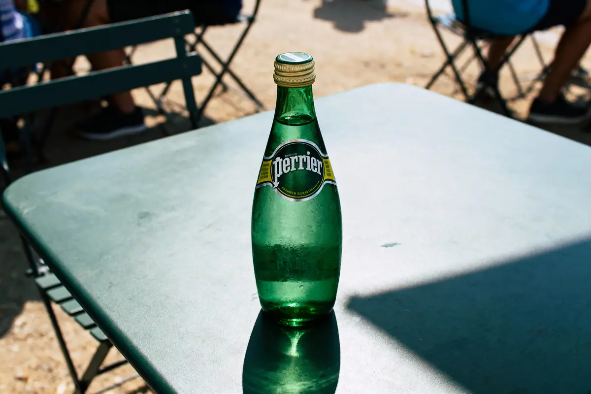 Bouteille verte de Perrier sur une table en métal, avec des sièges flous en arrière-plan extérieur.