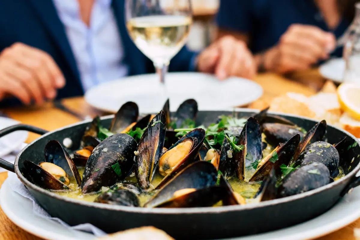 Plat de moules servi avec sauce, persil et verre de vin blanc en arrière-plan, ambiance de repas convivial.