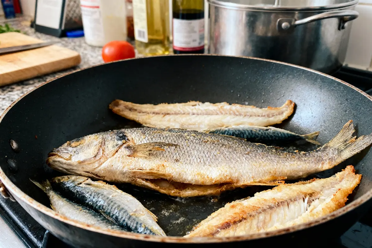 Poisson entier et filets cuisinés dans poêle noire sur cuisinière, cuisine en arrière-plan avec ustensiles.