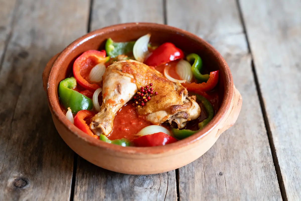 Plat en terre cuite avec poulet, poivrons, oignons et sauce tomate sur table en bois.
