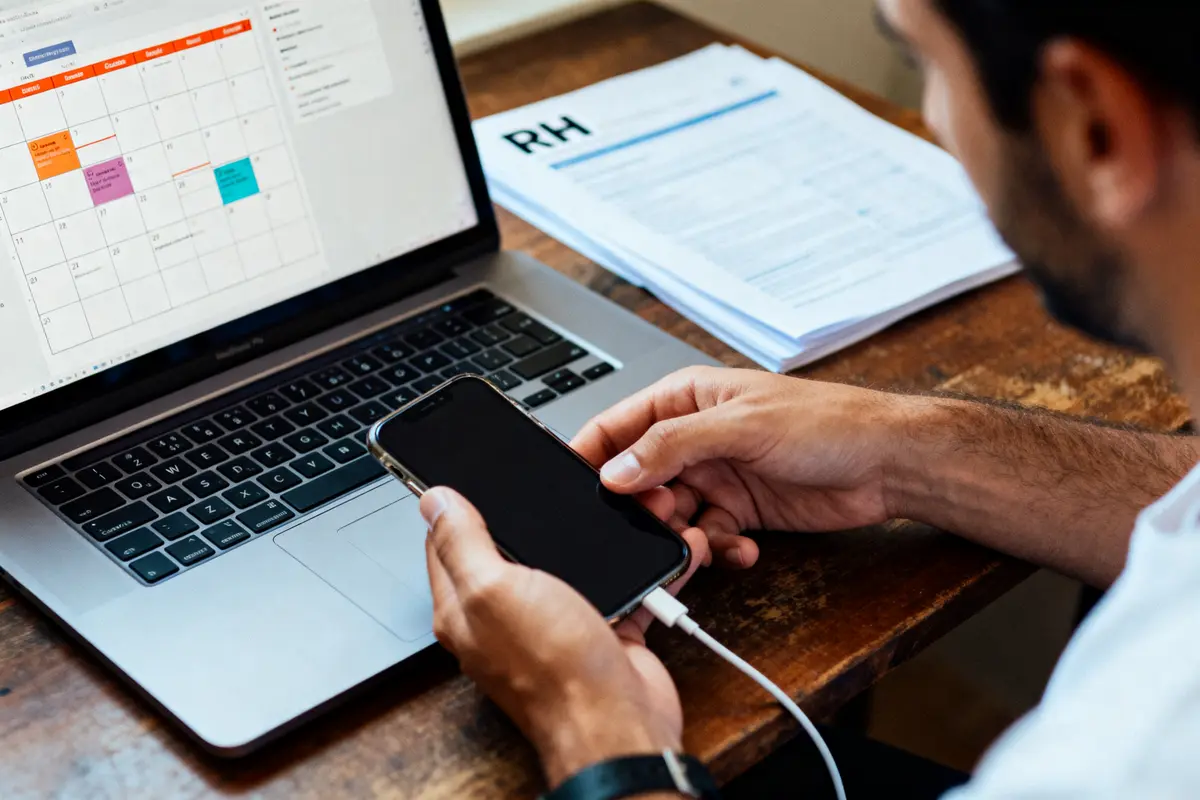 Homme consultant un calendrier électronique sur ordinateur portable et smartphone, documents RH sur la table en bois.