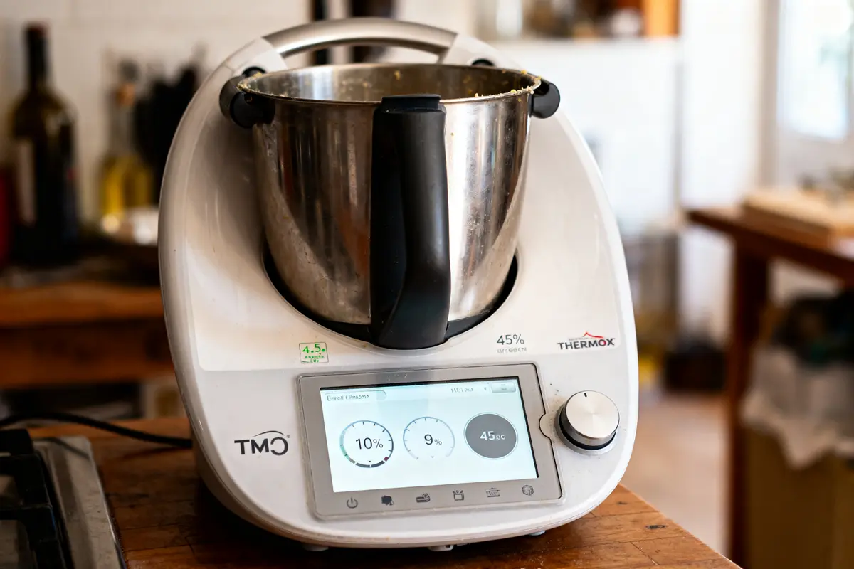 Robot culinaire Thermomix avec écran digital allumé dans une cuisine. Bol en acier inoxydable sur le dessus.