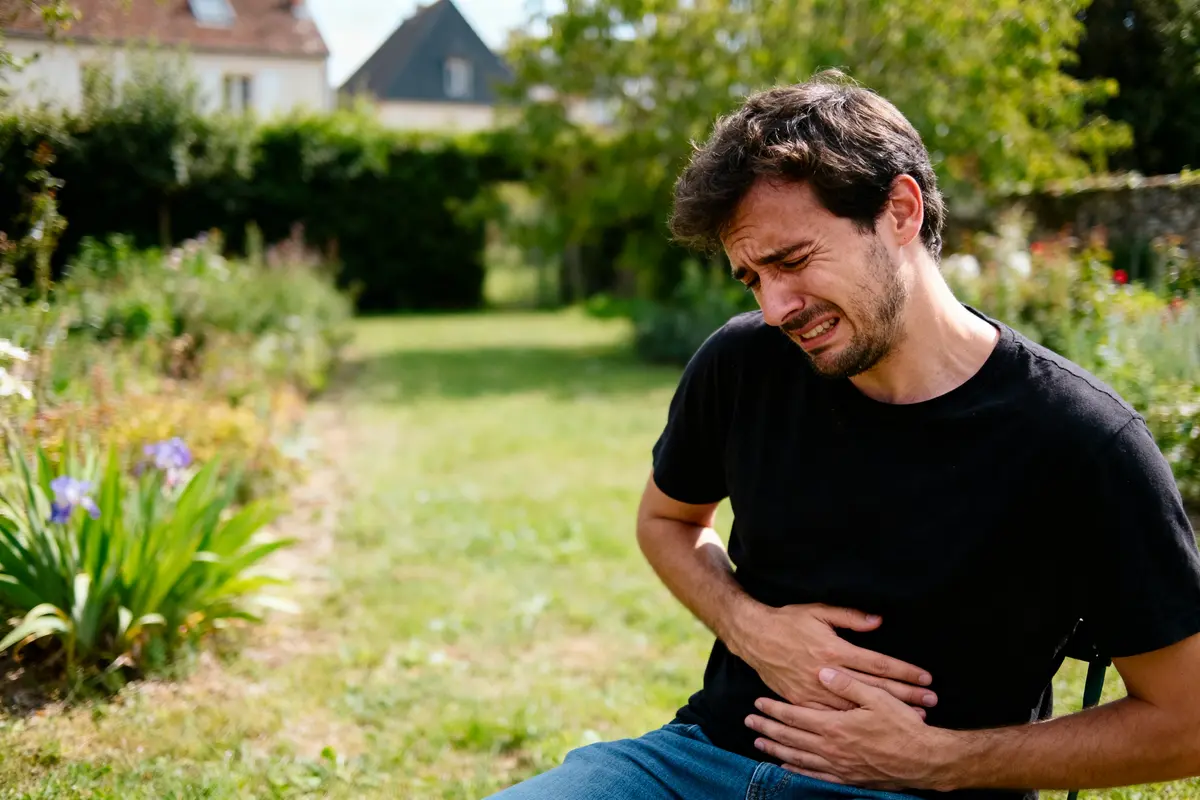 Homme en plein air souffrant de douleur abdominale, évocateur d'inconfort ou malaise, dans un jardin ensoleillé.