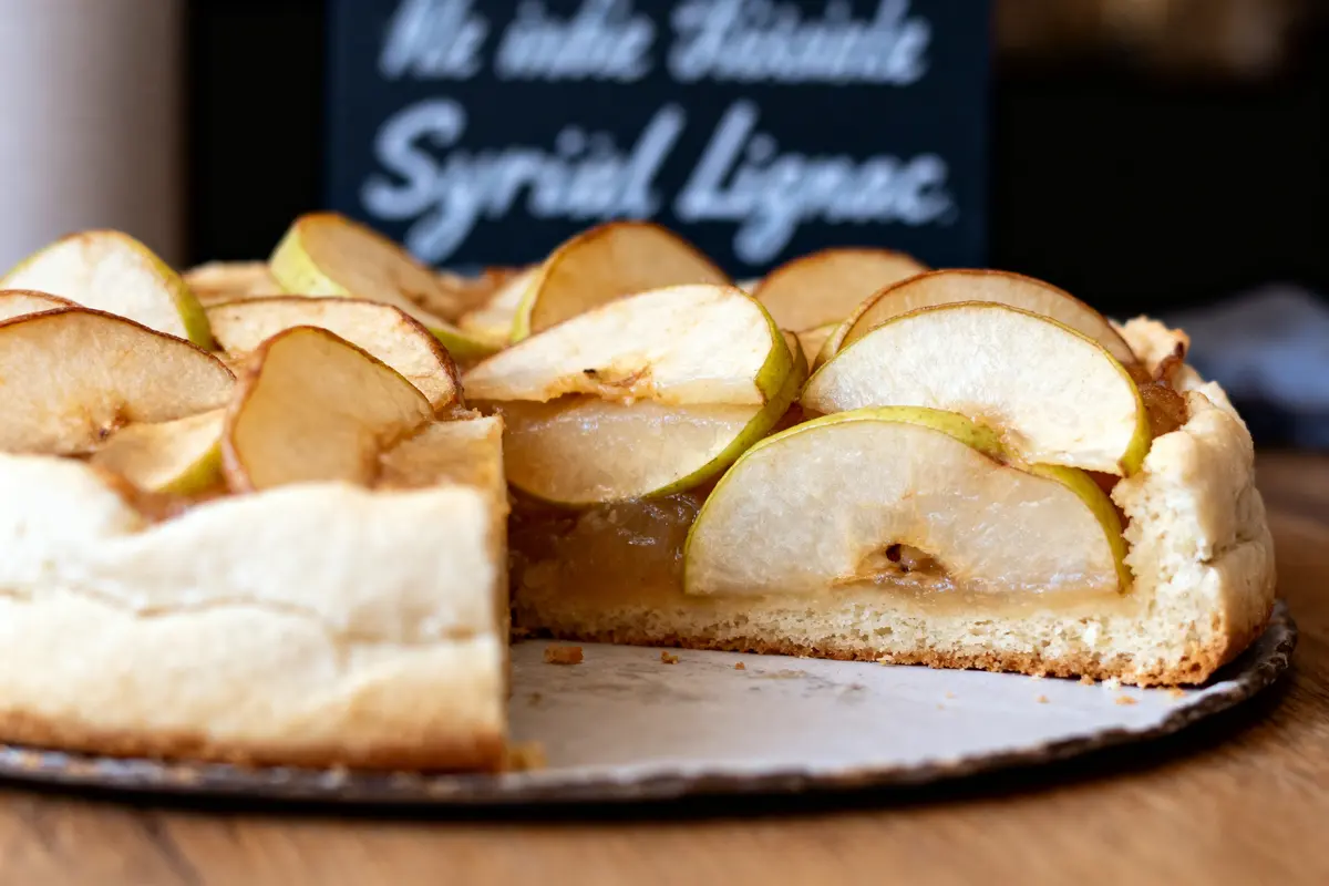 Gâteau aux pommes partiellement coupé sur une assiette, fond flou avec panneau écrit à la craie.